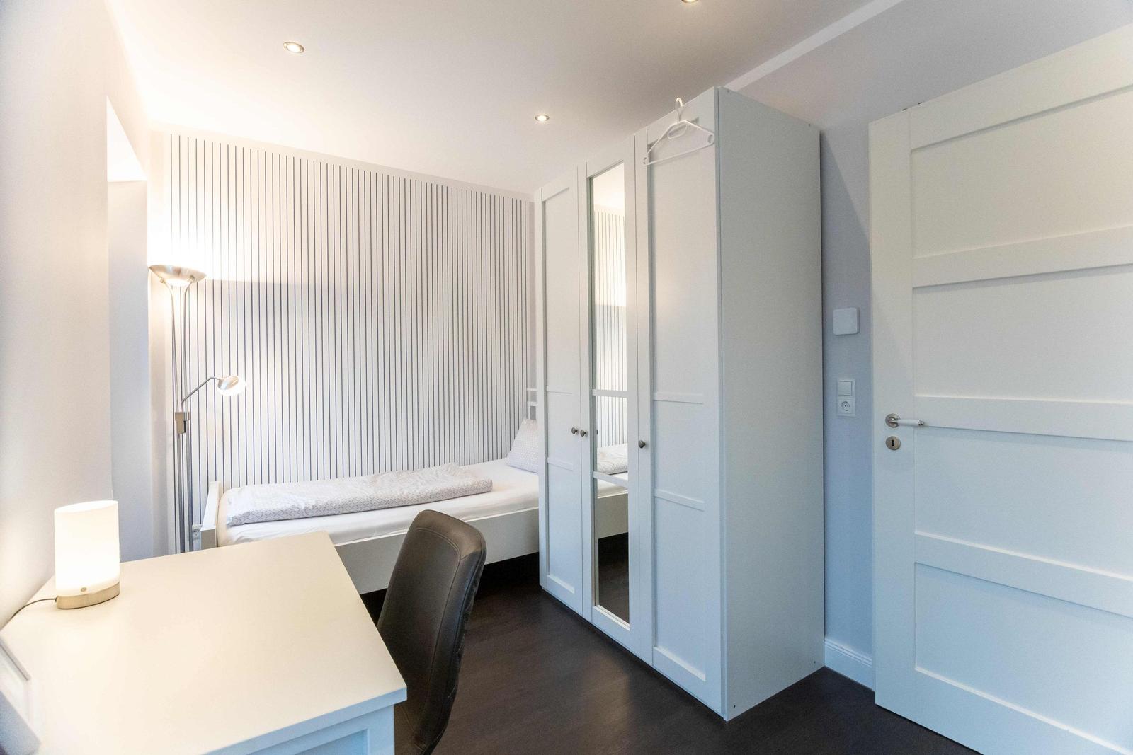 Schlafzimmer mit Bett, Nachttisch und Stehlampe. Fenster mit floralen Vorhängen.