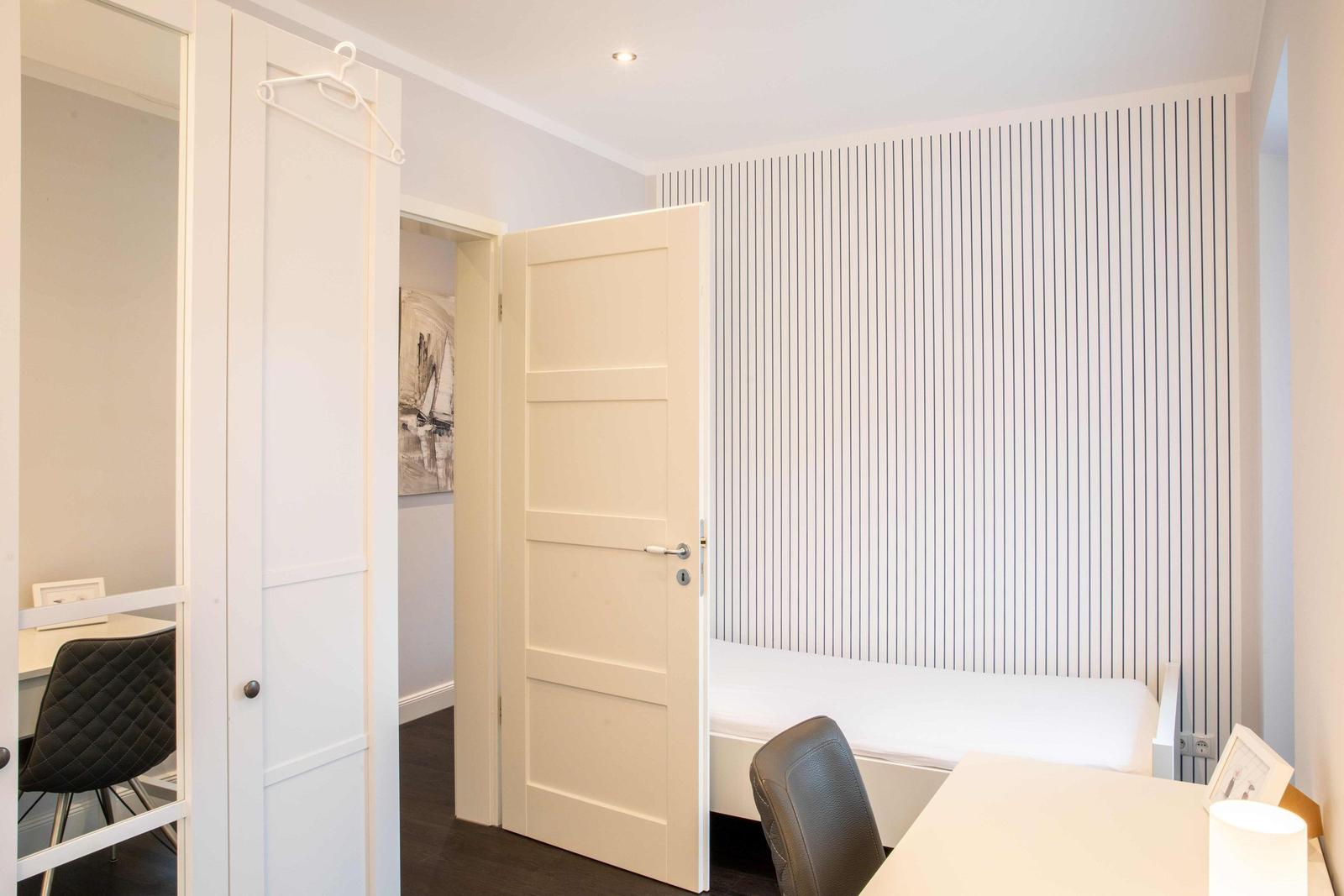 Schlafzimmer mit Bett, Schreibtisch und Kleiderschrank. Wand mit vertikalen Streifen.