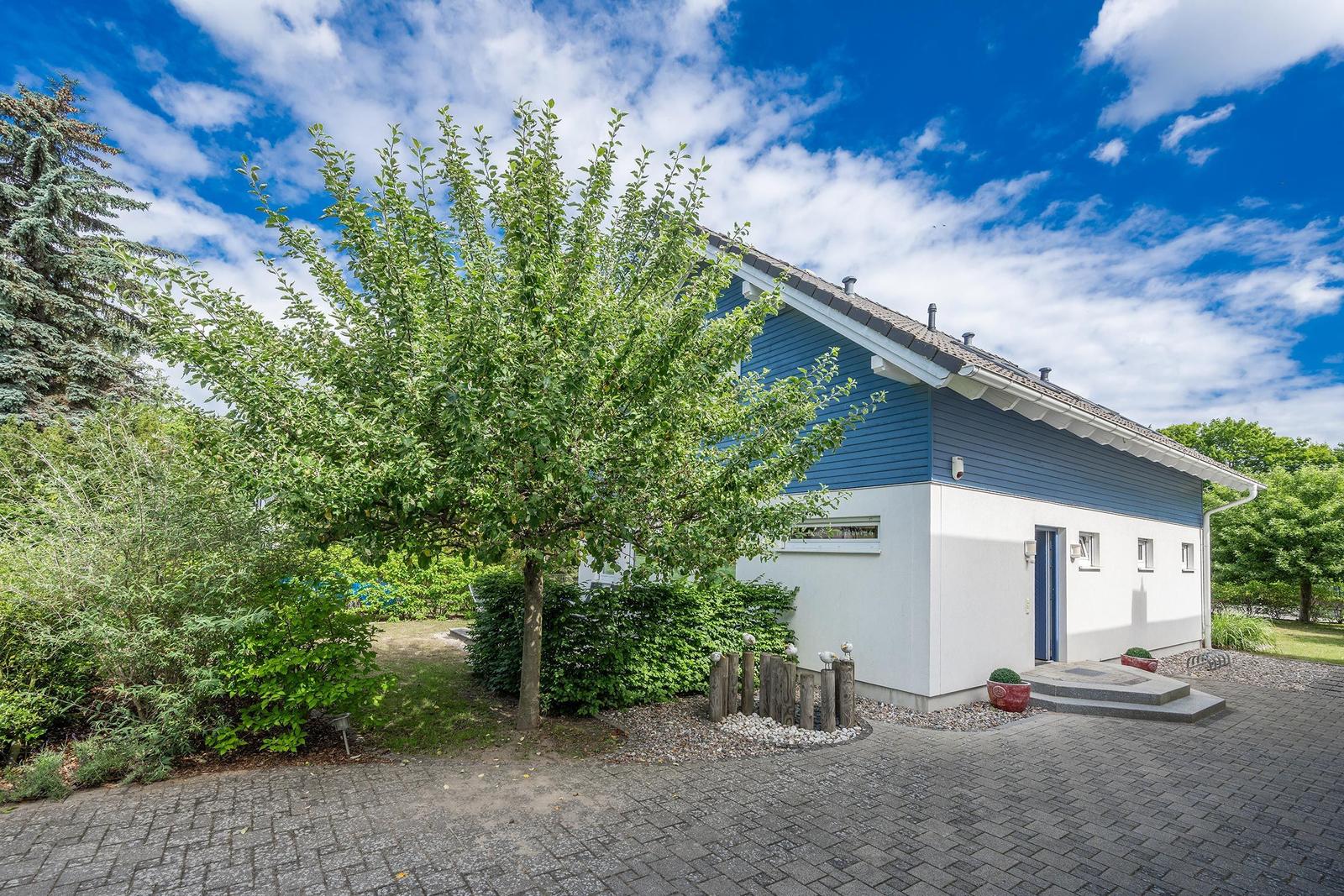 Blau-weißes Haus mit Garten und Terrasse. Gartenzaun und kleine Bäume sichtbar.