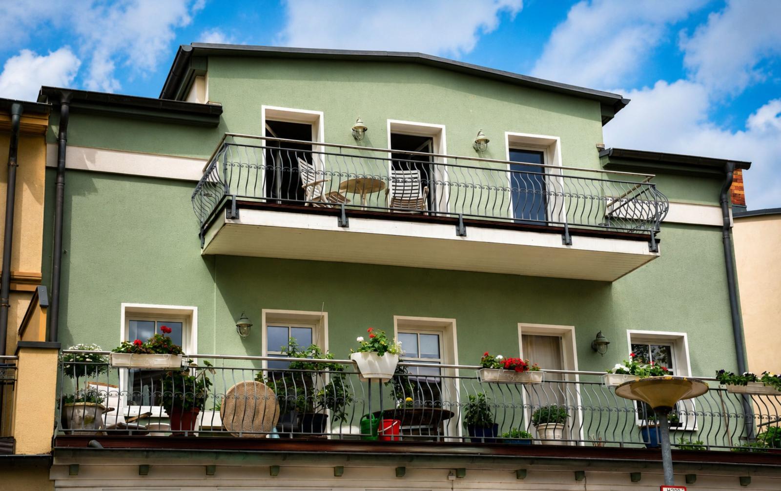 Grünes Gebäude mit Balkon und Fenstern. Unter dem Balkon sind Pflanzen zu sehen.