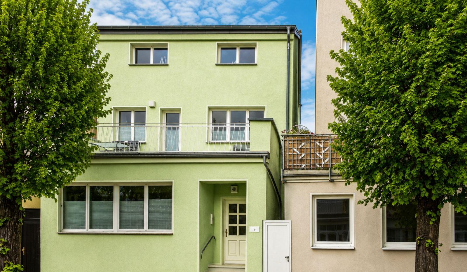 Grünes Mehrfamilienhaus mit Balkon und Eingangstür. Umgeben von Bäumen unter blauem Himmel.