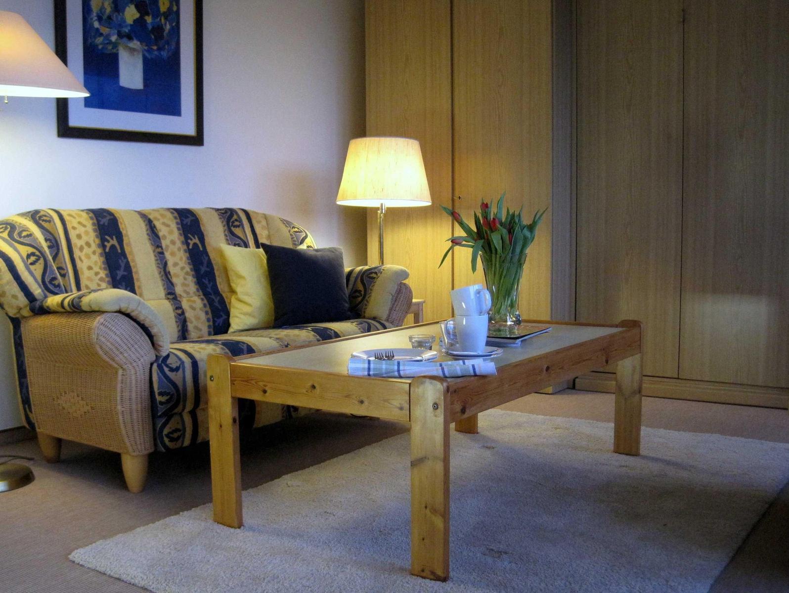 Ein Wohnzimmer mit Sofa, Tisch und Lampen. Auf dem Tisch stehen Tassen und Blumen.