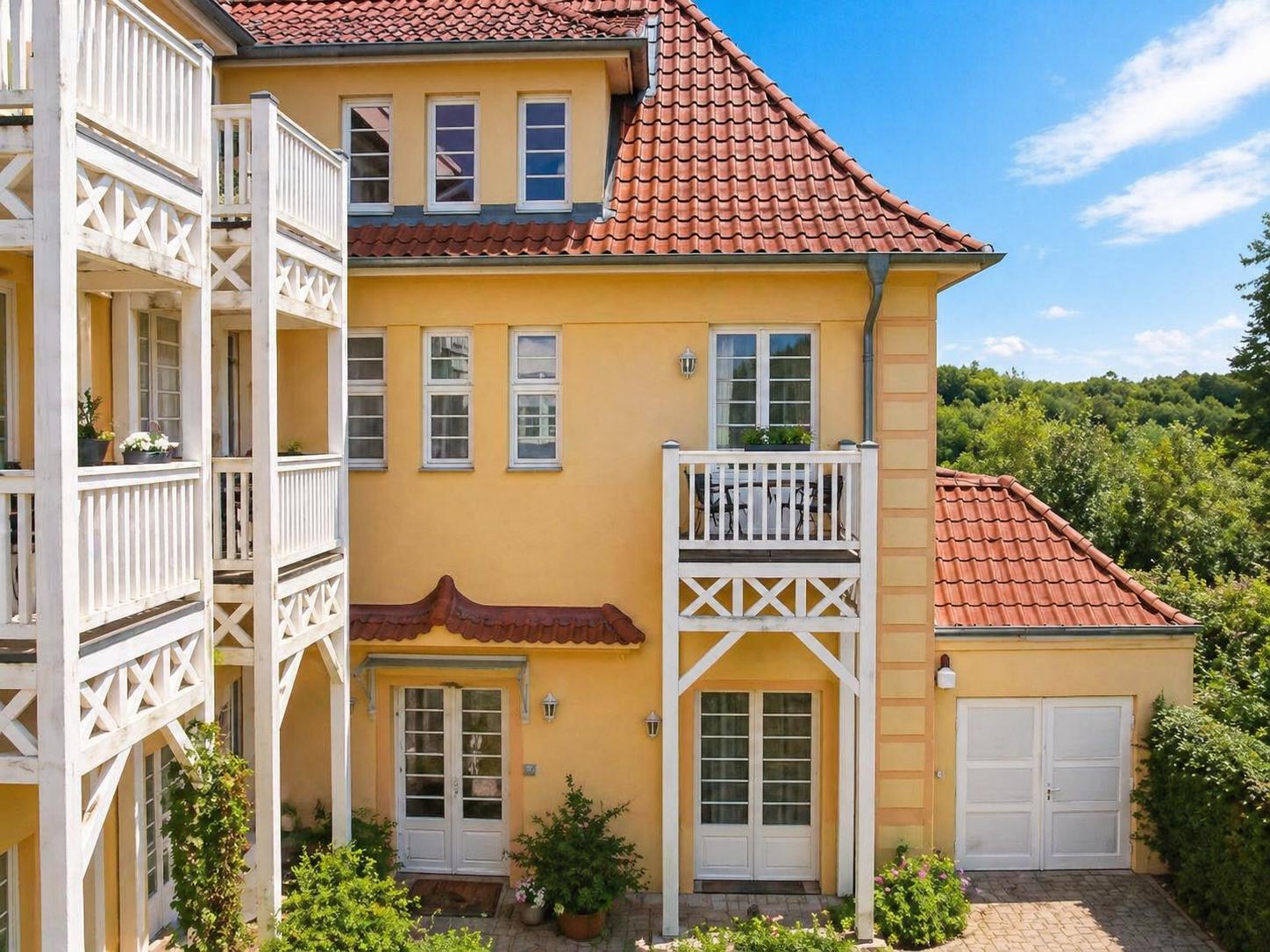 Ein gelbes Haus mit rotem Dach und weißen Balkonen. Umgeben von Grün und einem Garagenbereich.