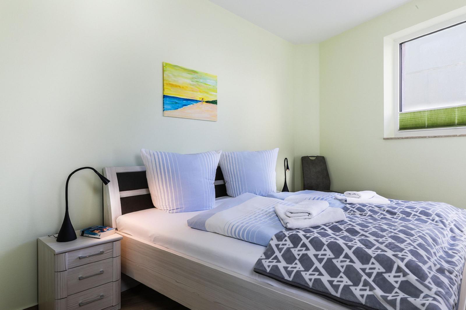 Ein Schlafzimmer mit Bett, Tisch und Stühlen. An der Wand hängen Bilder. Durch das Fenster ist ein Balkon sichtbar.