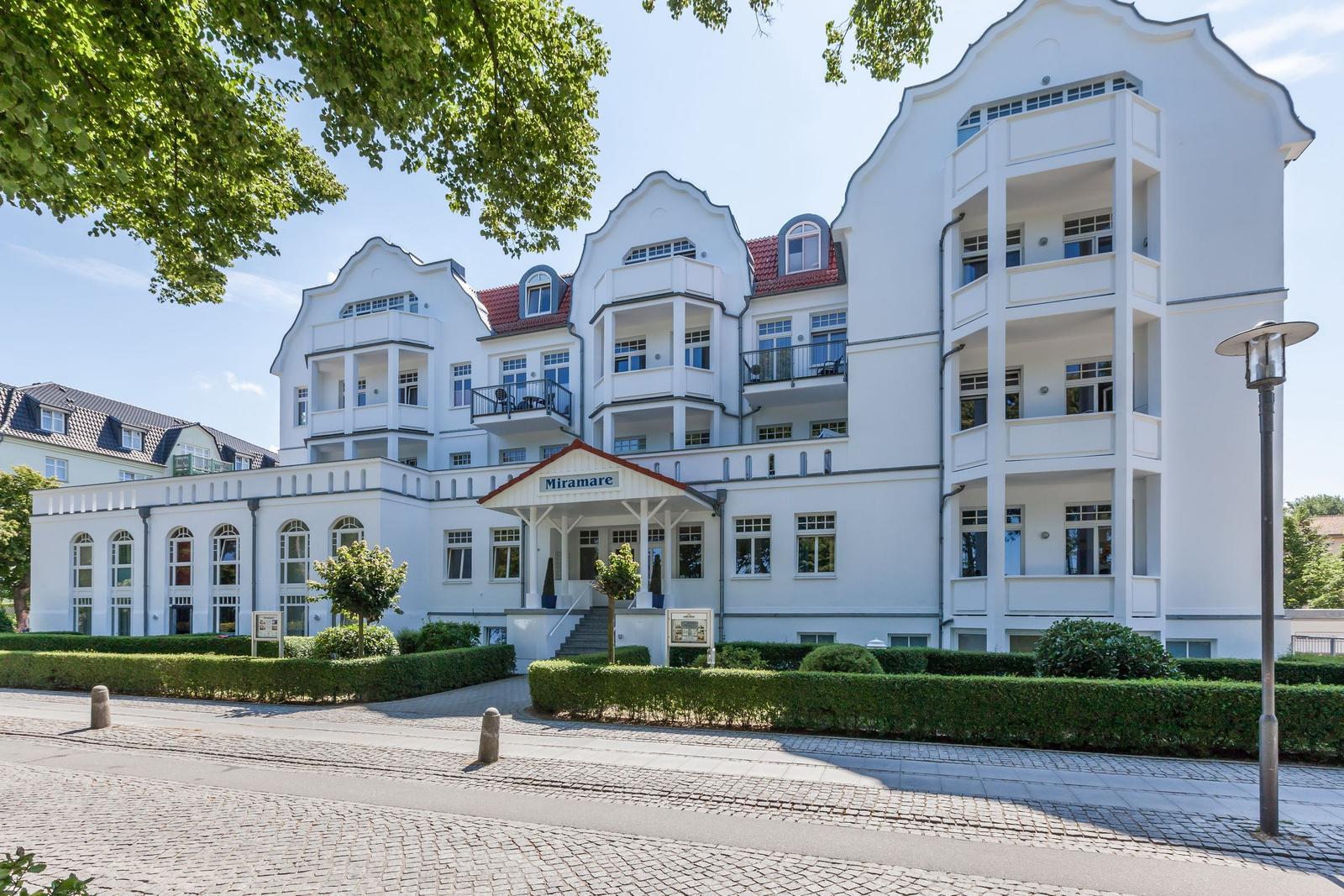 Weißes Mehrfamilienhaus mit Balkonen und rotem Dach unter Bäumen.