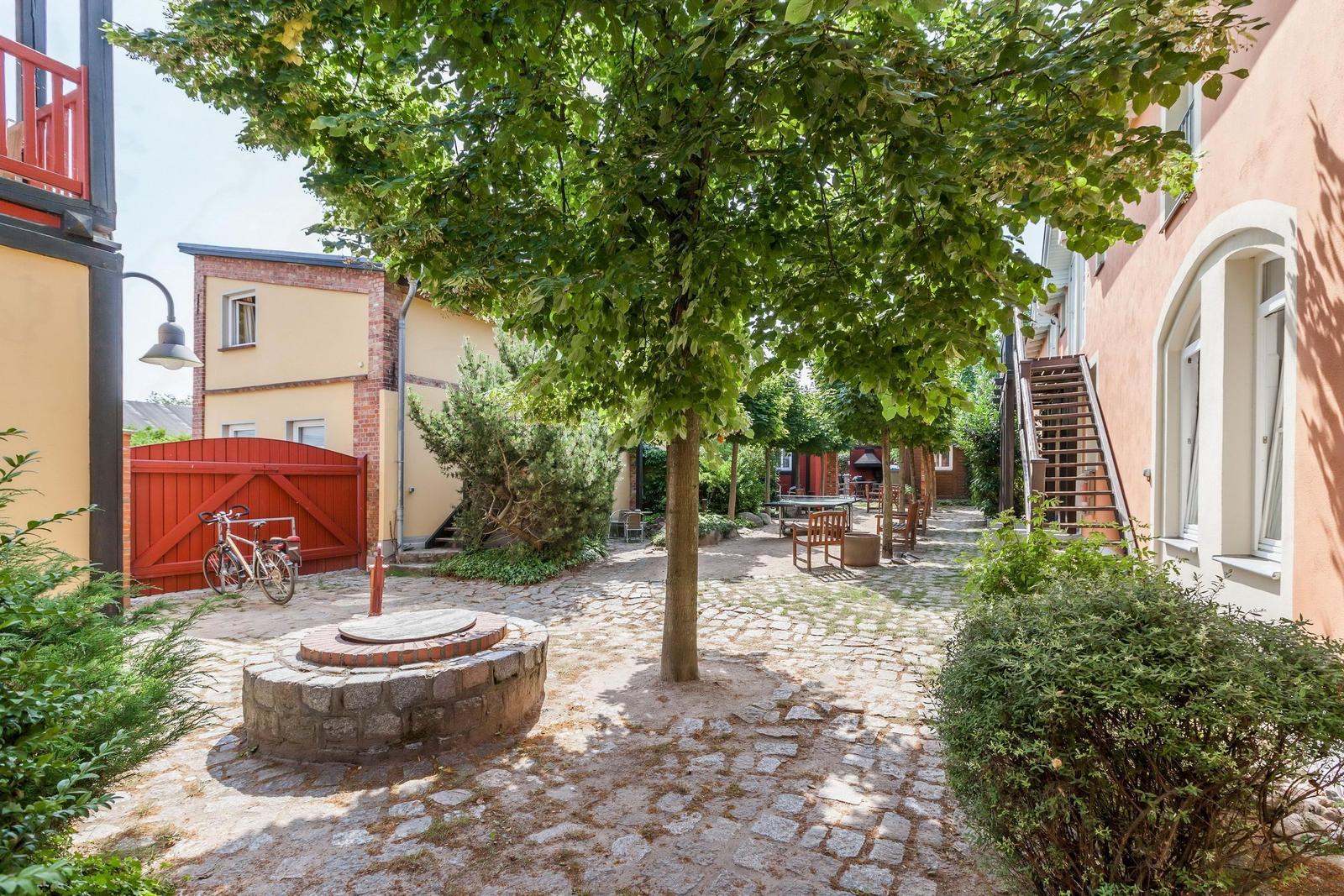 Kiesweg durch einen gepflegten Innenhof mit Bäumen, Sitzgelegenheiten und einem Brunnen.