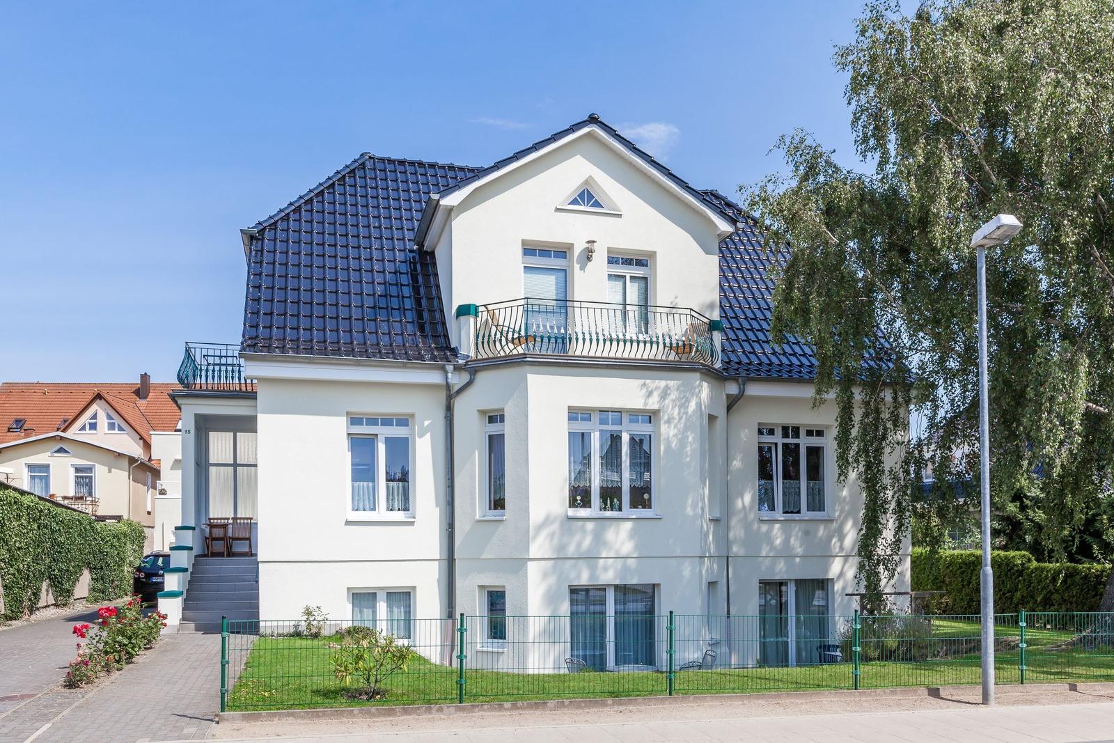 Weiße Villa mit Balkon und grünem Garten. Schwarzes Dach und Fenster mit Vorhängen.