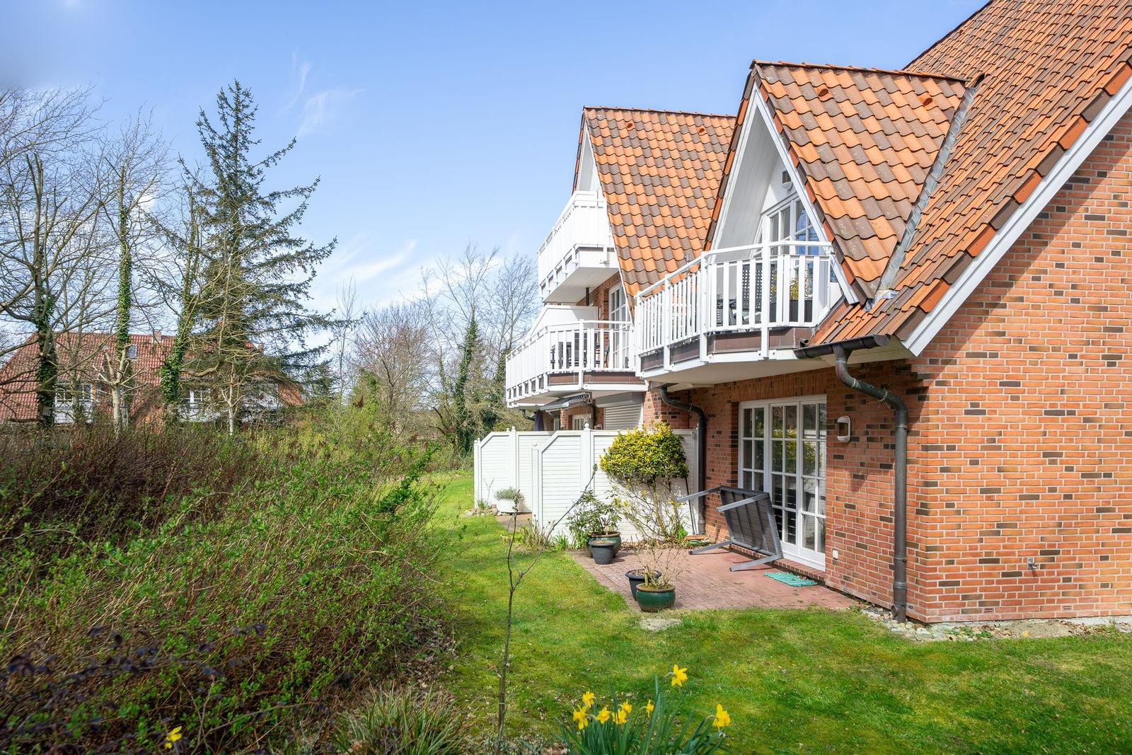 Haus mit rotem Ziegeldach, weißen Balkonen und Glasfenstern. Vorgarten mit Gras und Pflanzen.