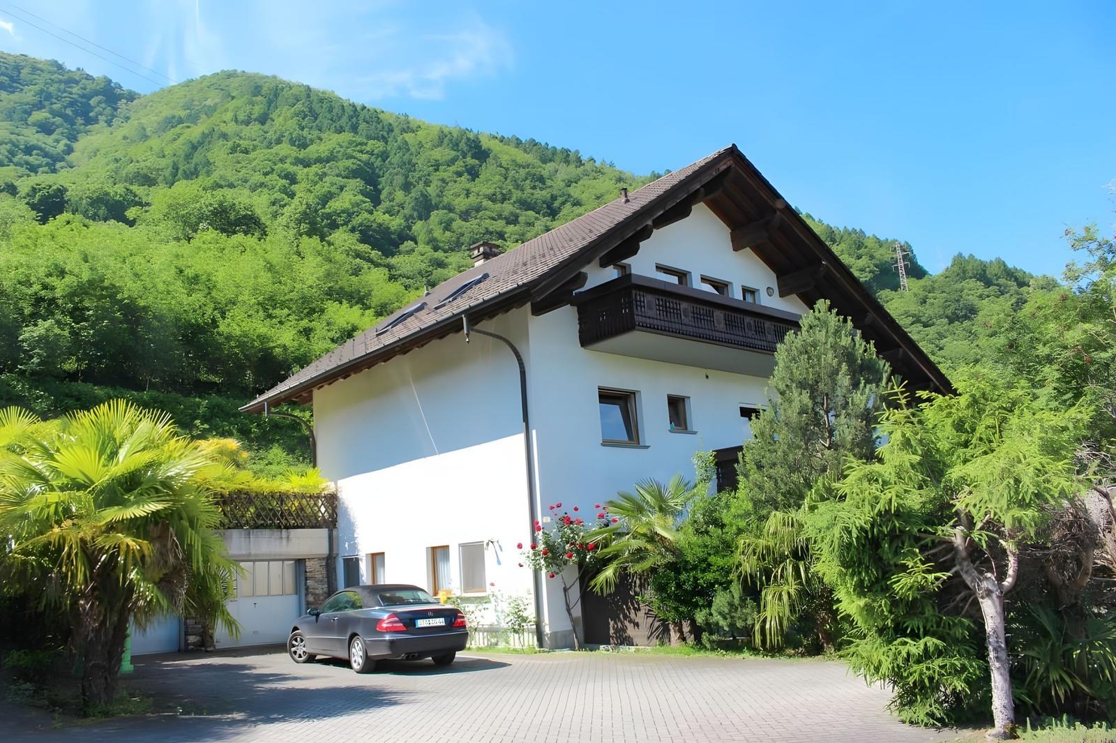 A two-story house with a balcony, surrounded by trees and mountains. A car is parked in front of the house.