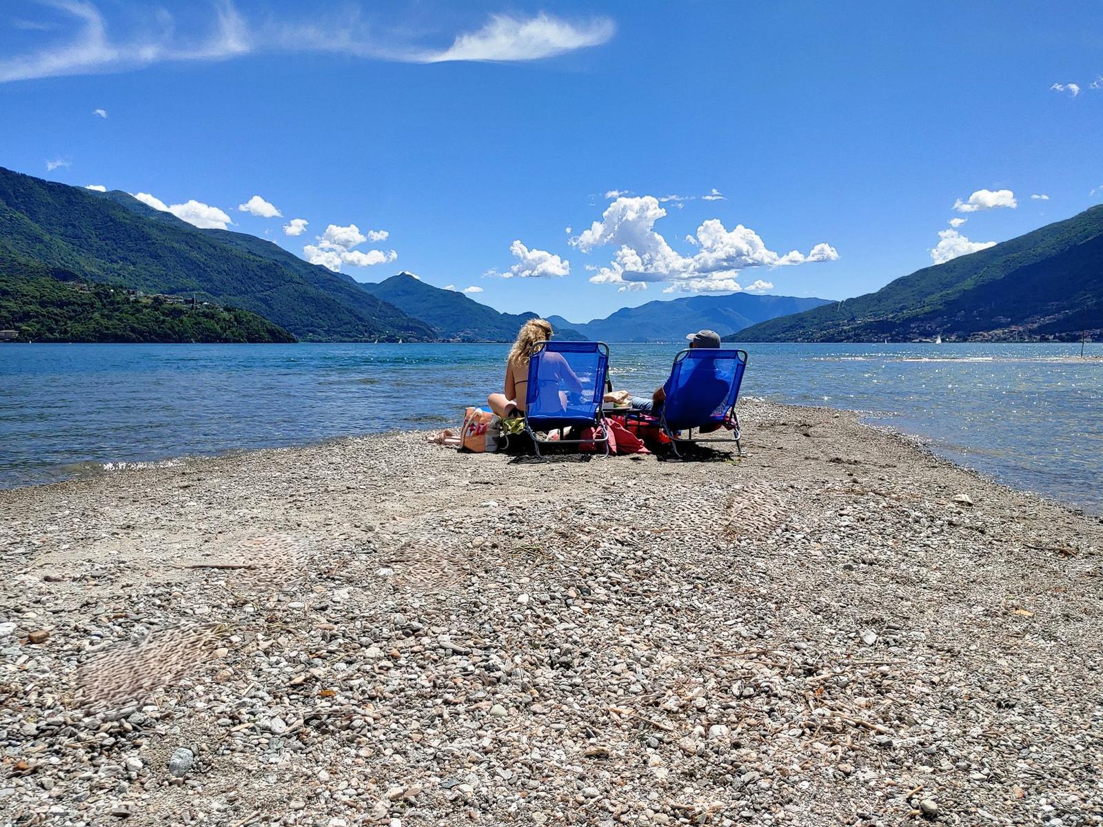 Terrasse mit Sonnenschirmen am See, blauer Himmel und Berge im Hintergrund.