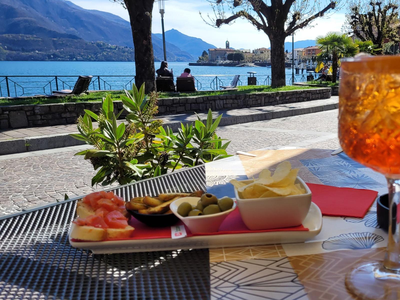 Zwei Personen sitzen in blauen Liegestühlen am steinigen Ufer eines Sees mit Bergen im Hintergrund.