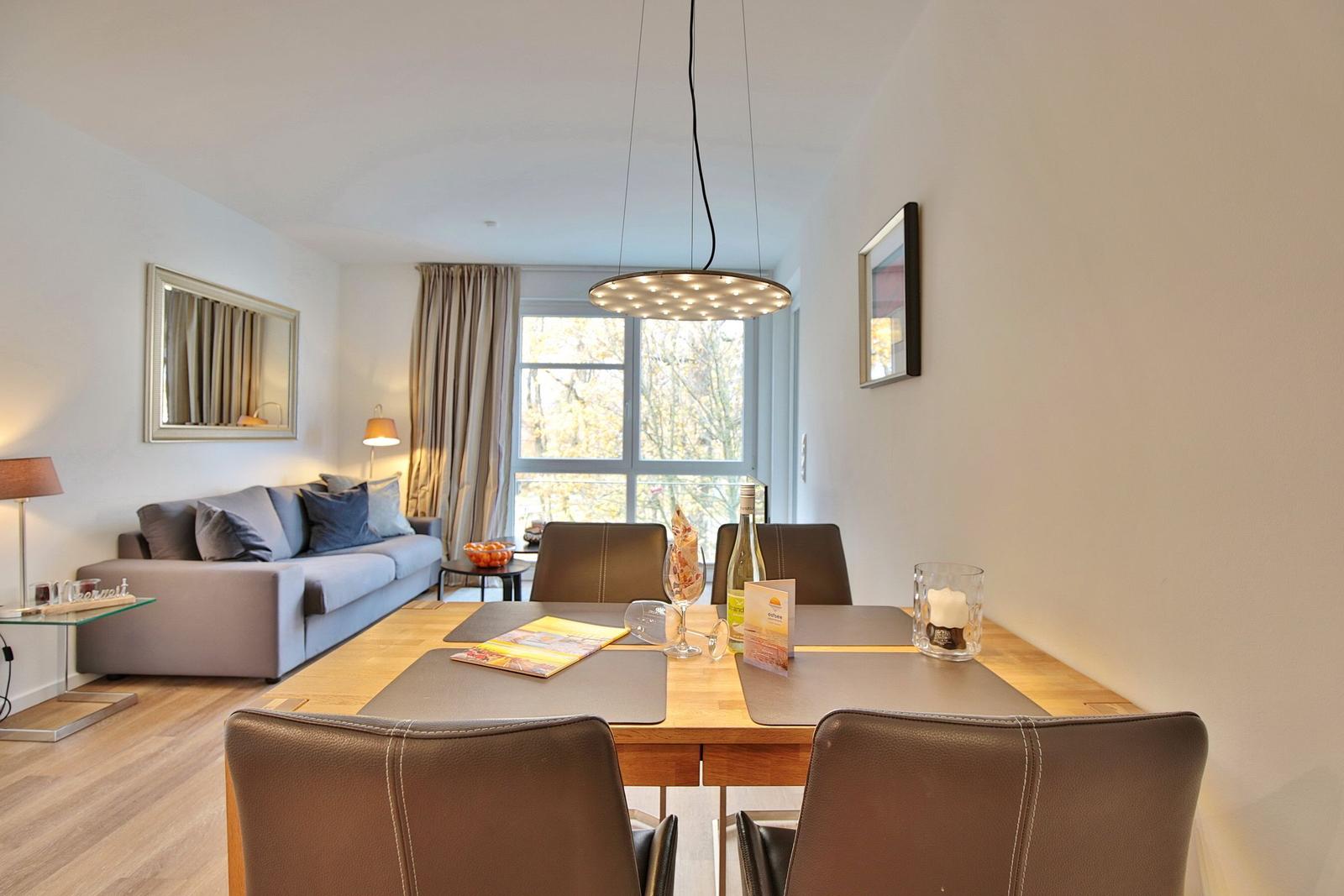 Essbereich mit Tisch und Stühlen. Im Hintergrund Wohnbereich mit Sofa und Fenster.