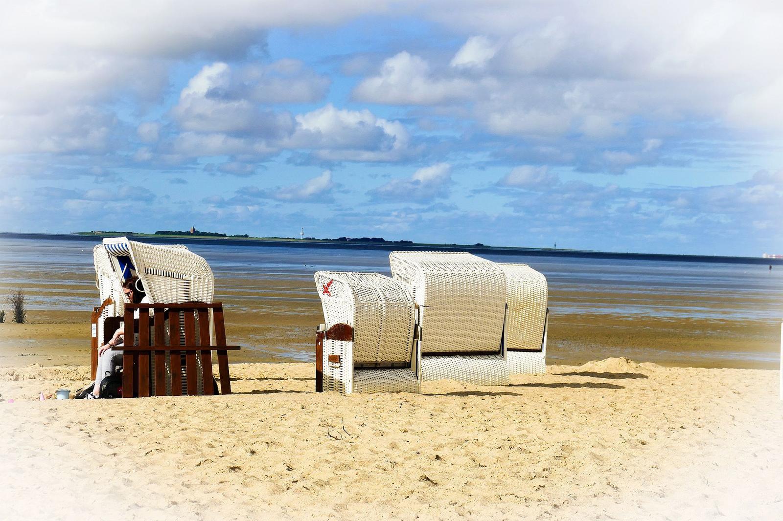 Two white beach chairs stand on the sandy shore. Water stretches into the distance under a cloudy sky.