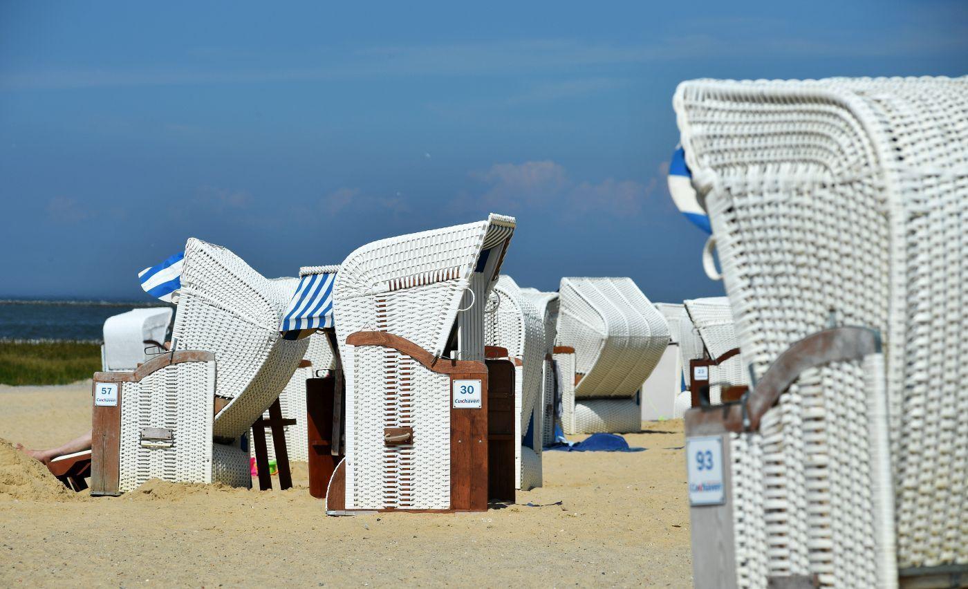 Zwei weiße Strandkörbe stehen auf Sand, im Hintergrund das Meer und Badegäste.