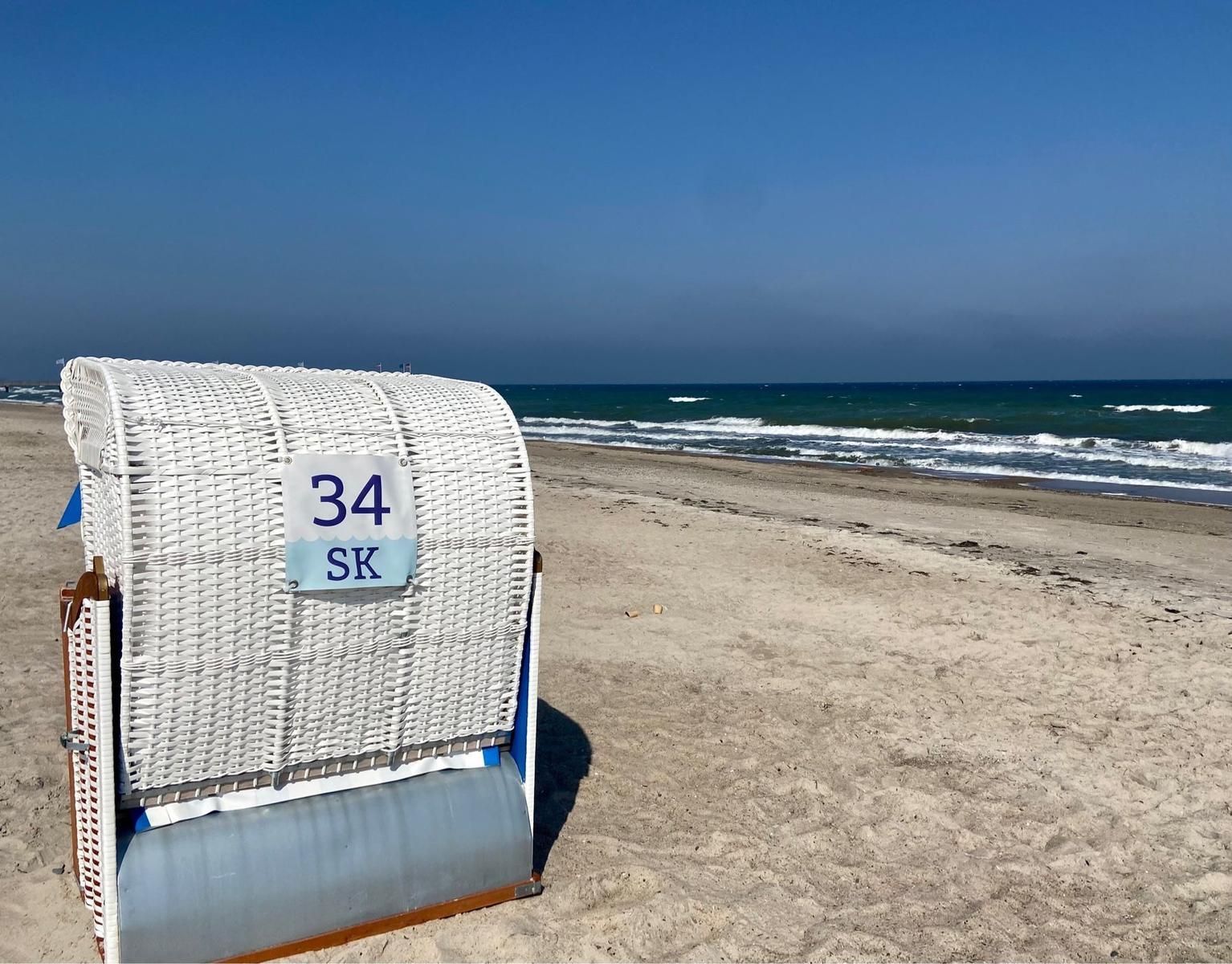 Weißer Strandkorb mit der Nummer 34 SK auf sandigem Strand vor dem Meer.