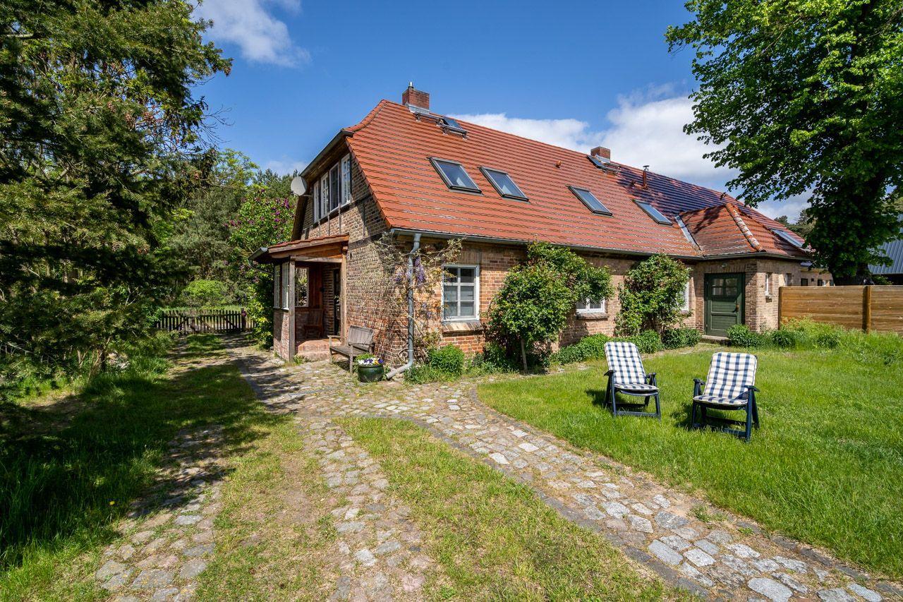 Green garden with table, chairs, and umbrella. Stone path and trees in background.