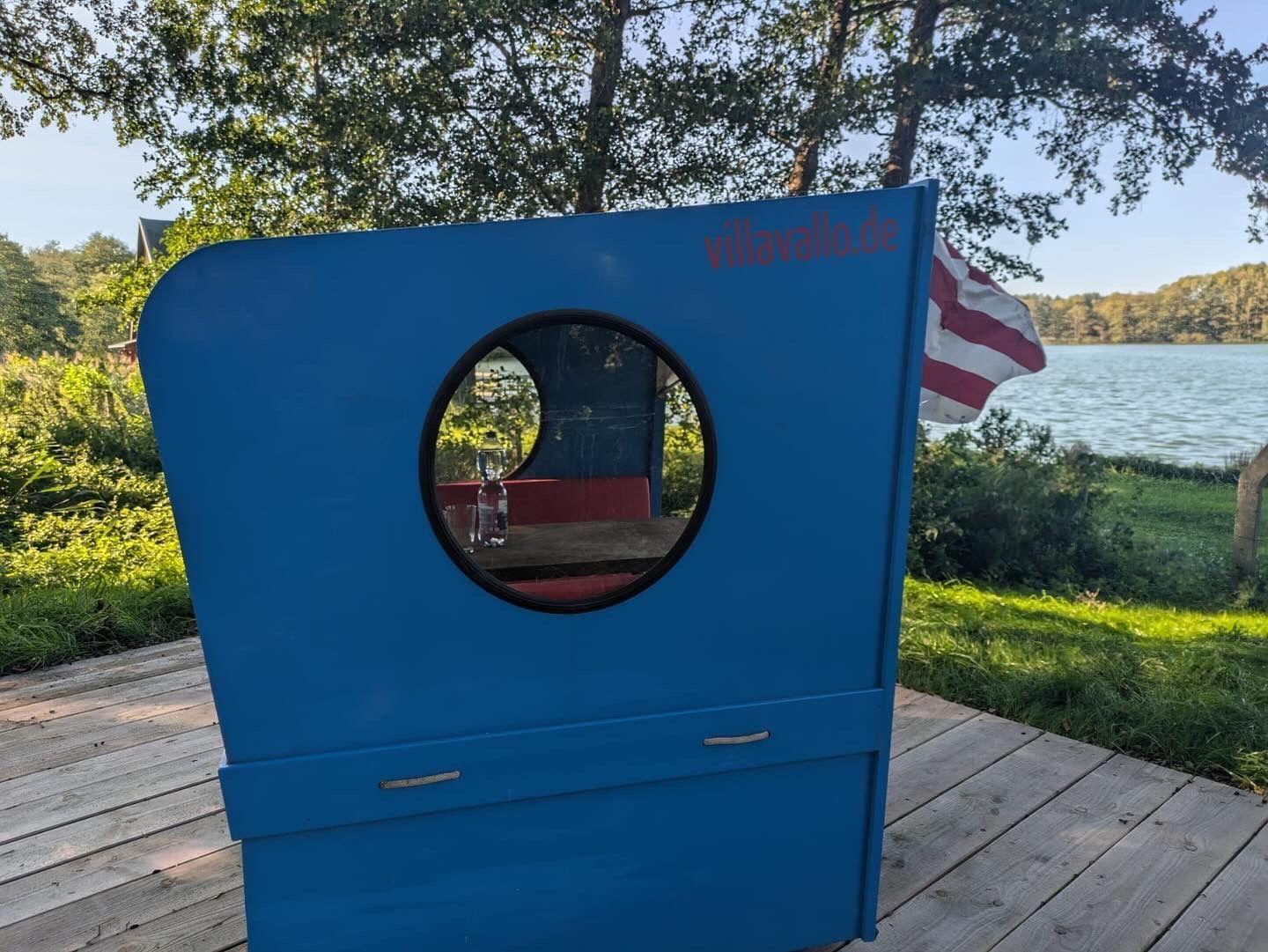 Ein blauer Pavillon mit rotem Sitzbereich steht am Ufer eines Sees. Im Vordergrund brennt ein Feuer.