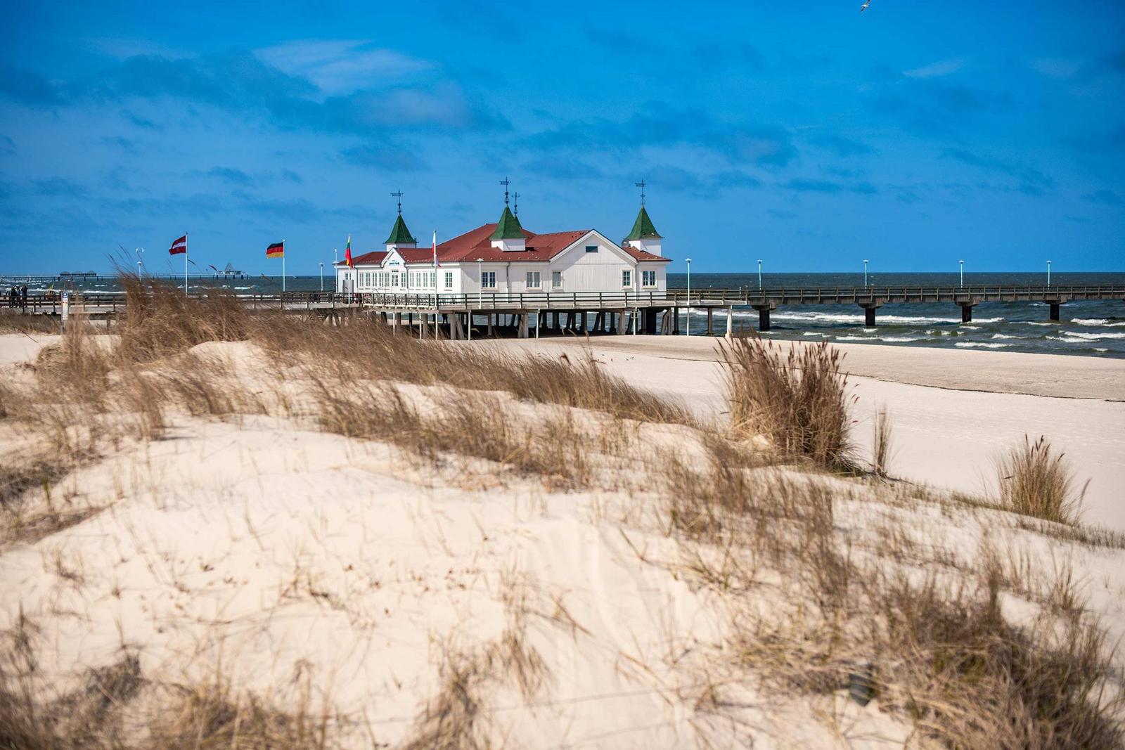 Strand mit weißen Strandkörben, Steg und Haus im Hintergrund bei Sonnenuntergang.
