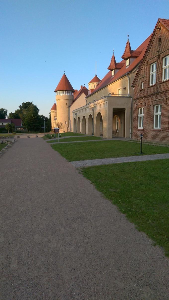 Schloss mit Turm, rotem Dach und Fenstern, umgeben von Bäumen und Blumen.