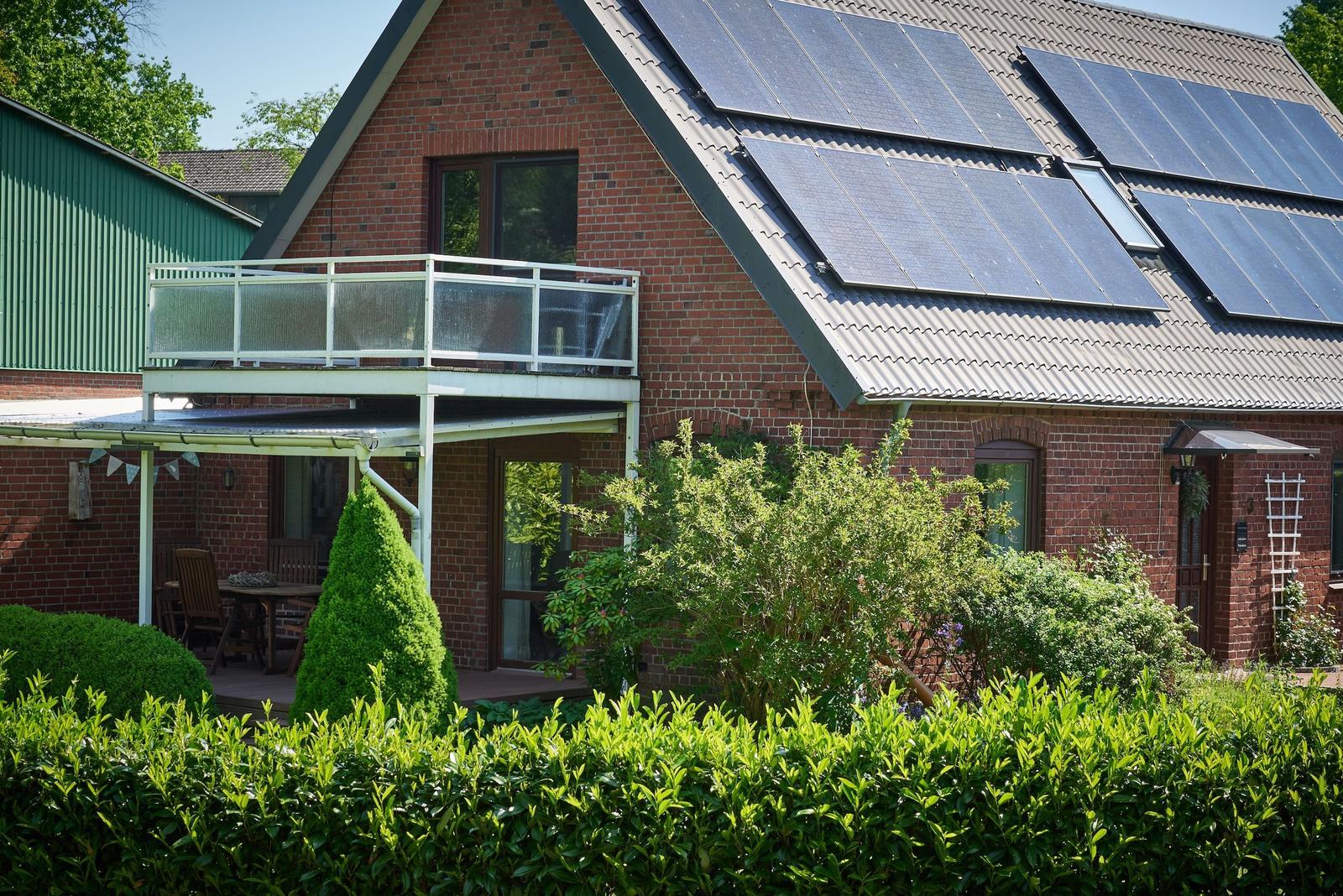 Red brick house with dark roof, surrounded by trees and greenery. A trampoline in the foreground.