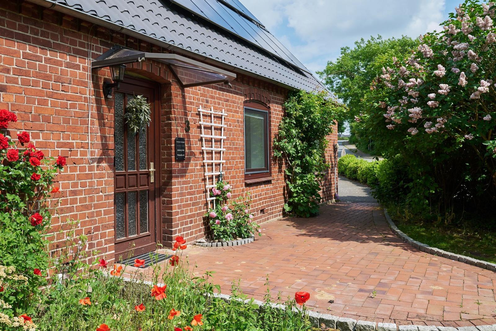 Red brick building with roof, windows, and door, surrounded by greenery and flowers.