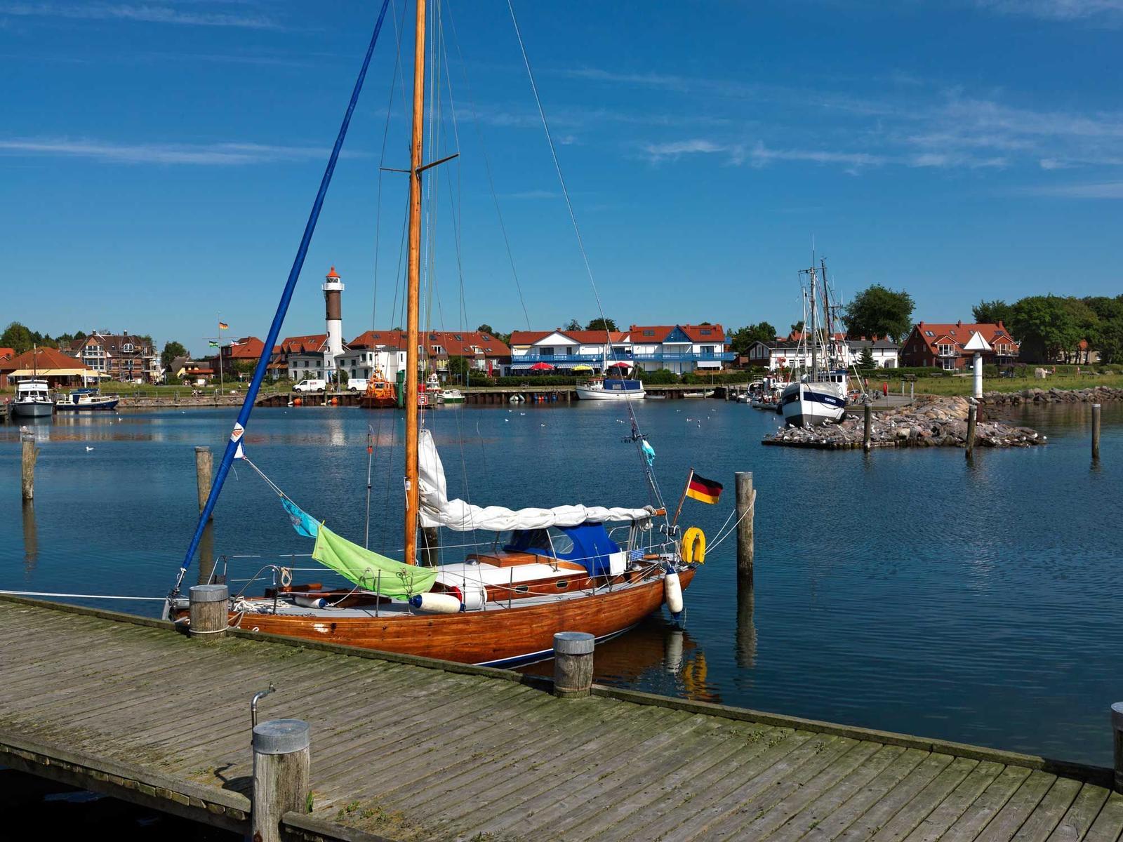 Holzboot liegt am Steg im Hafen. Im Hintergrund sind Häuser und Leuchtturm zu sehen.