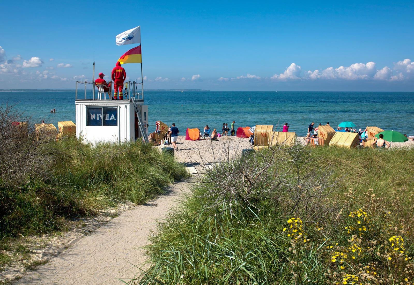 Strand mit Rettungswache, Strandkörben und Badegästen unter blauem Himmel.