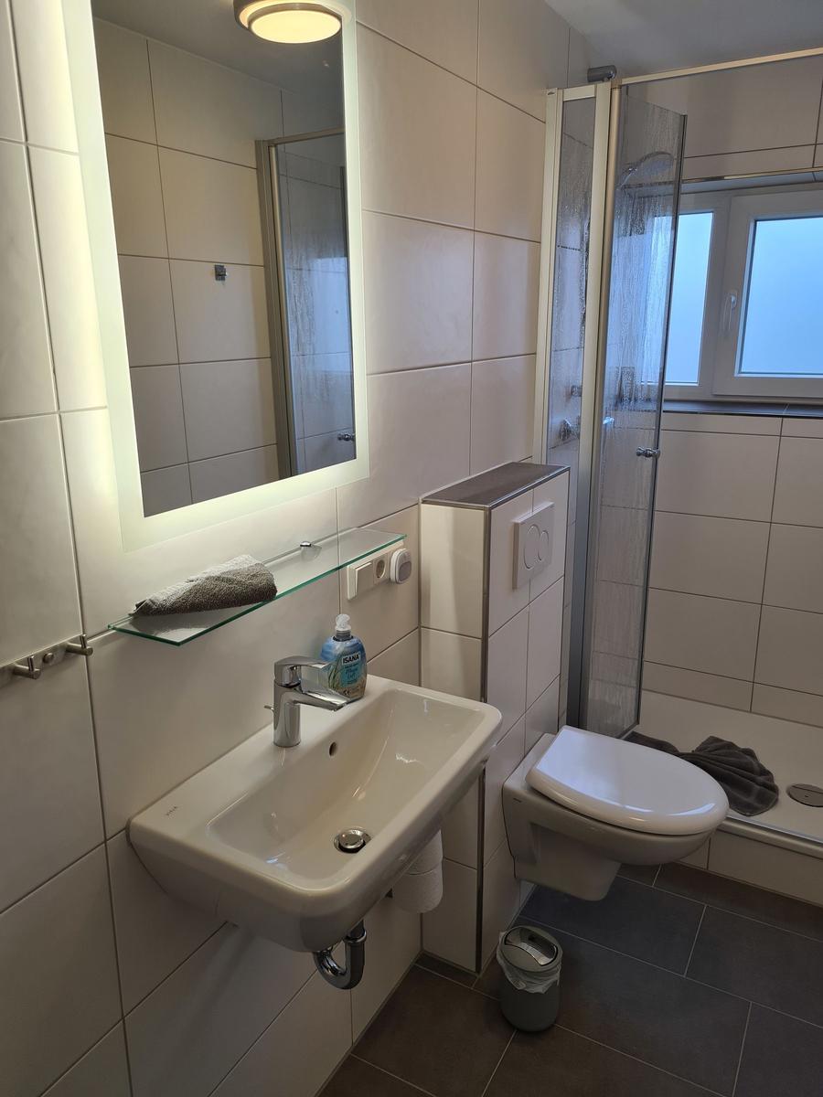 Badezimmer mit Dusche, Waschbecken und Toilette. Weiße Fliesen und graue Bodenfliesen.