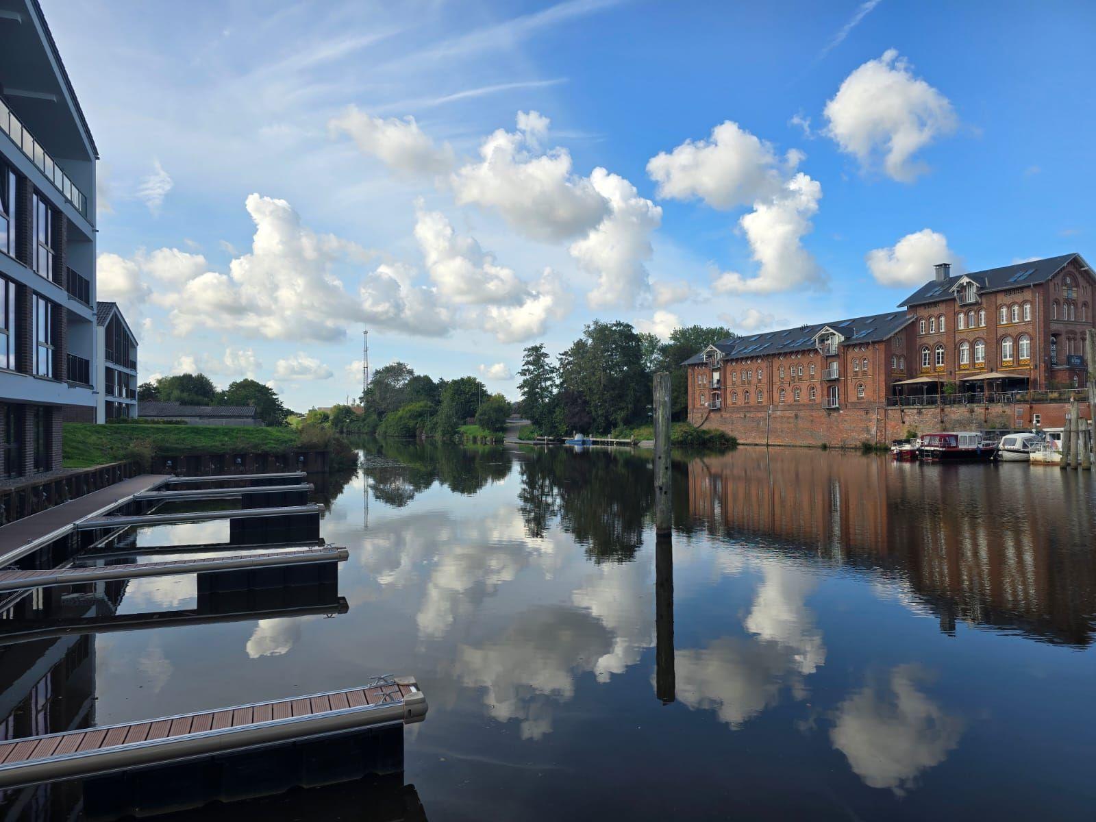 Hafen mit Steg, Booten und Reflexionen unter blauem Himmel mit weißen Wolken.