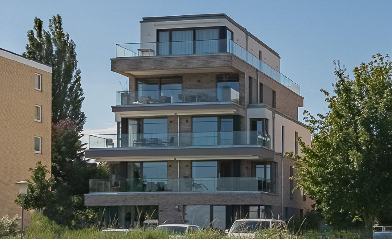 Modernes Mehrfamilienhaus mit Glasgeländern und Balkonen. Umgeben von Bäumen und Grünflächen.