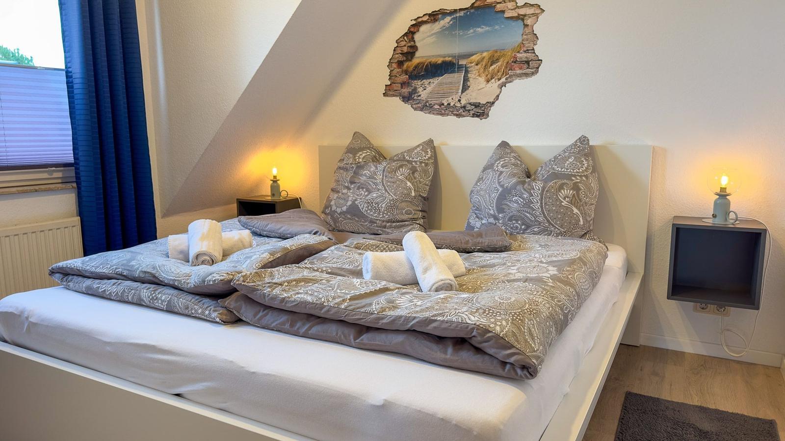 Bed with patterned duvet and pillows. Rolled towels on bed. Nightstand with lamp.