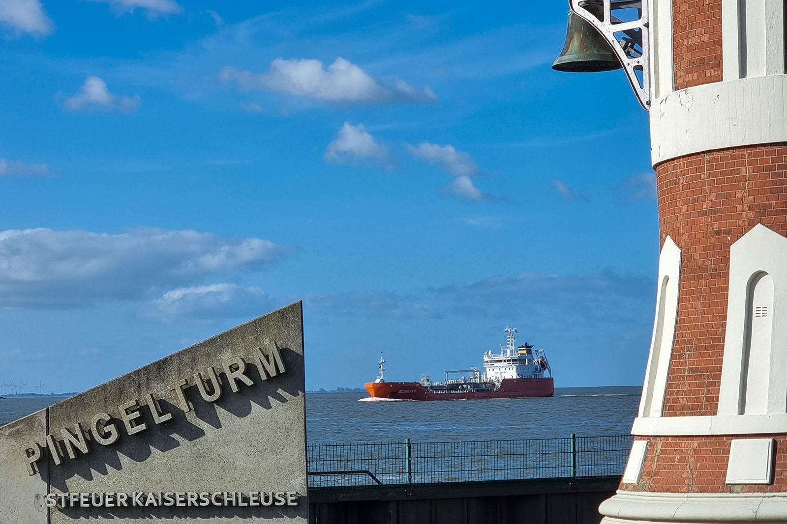 Rotes und weißes Leuchtturm-Objekt am Wasser mit Boot und Steinanlagen im Vordergrund.