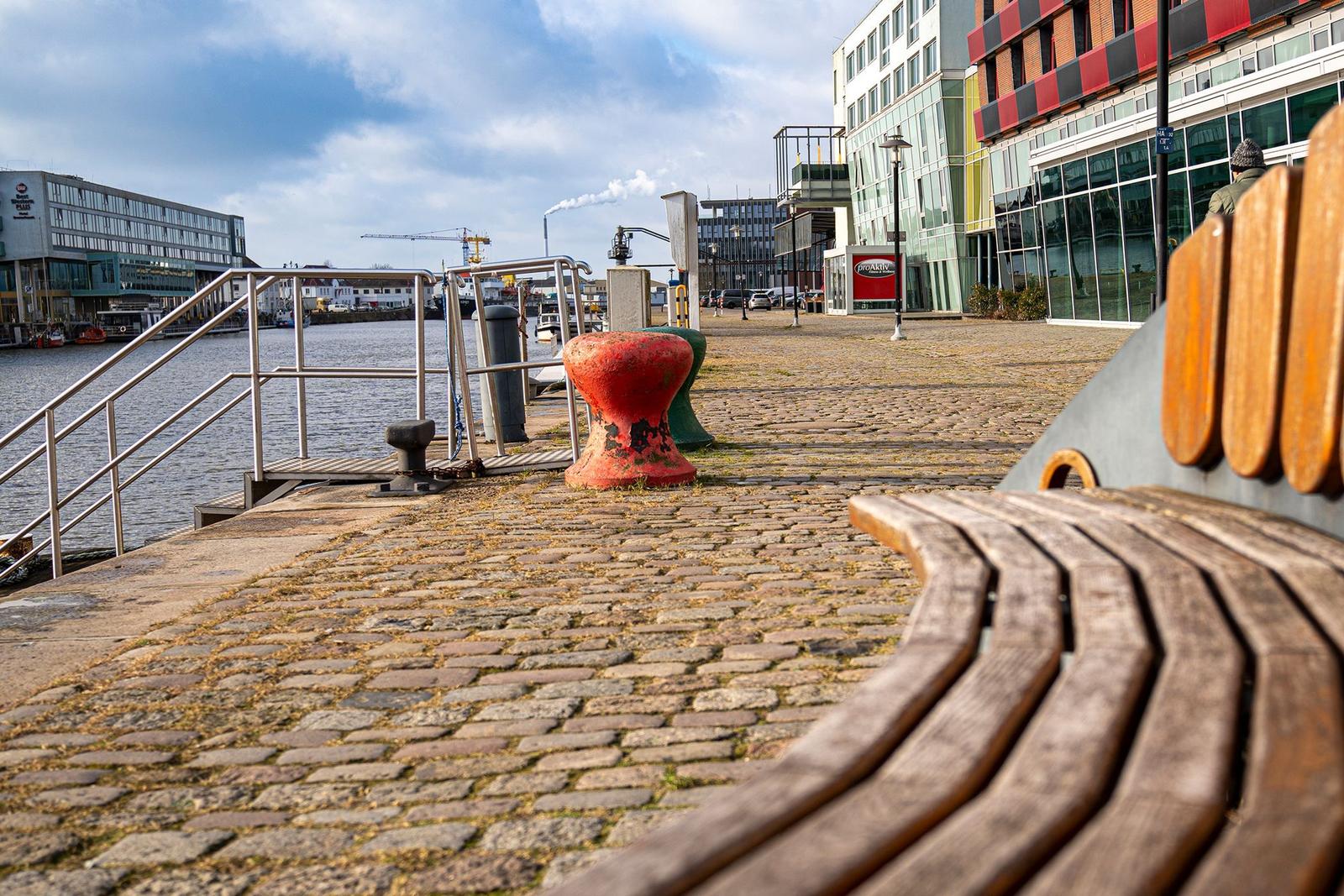 Rostiger Ankerpunkt am Hafen mit Booten und Gebäuden im Hintergrund.