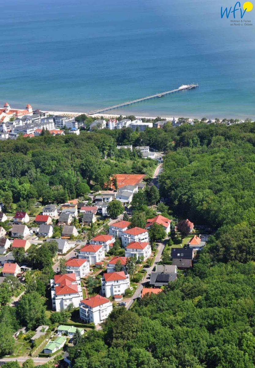 Aerial view of coastal town with red-roofed houses, greenery, and a pier extending into the sea.