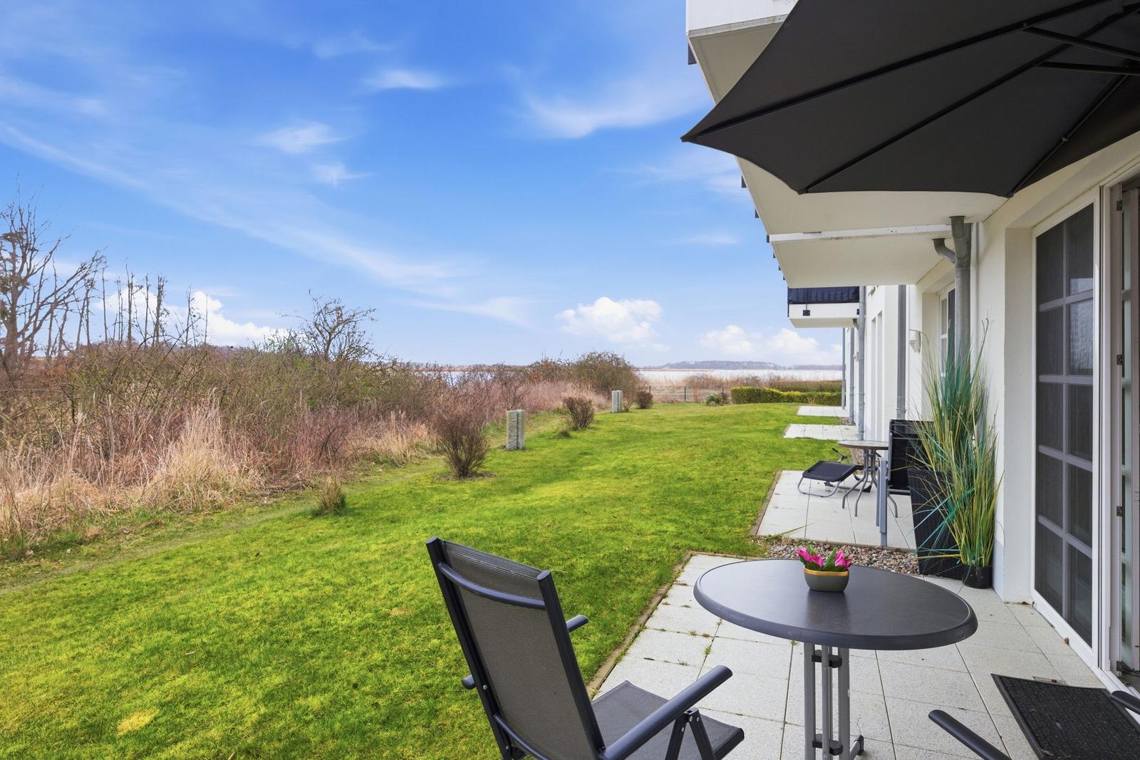 Terrasse mit Tisch, Stuhl und Sonnenschirm. Blick auf Grasfläche und Wasser.