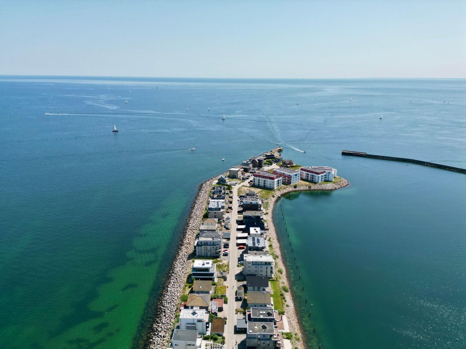 Strand mit klarem Wasser und Häusern im Hintergrund. Blick durch Vegetation.