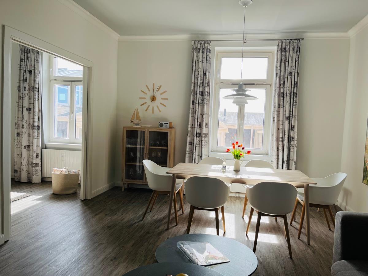 Wohn- und Essbereich mit Sofa, Tisch und Stühlen. Fenster mit Vorhängen. Wanddekoration.