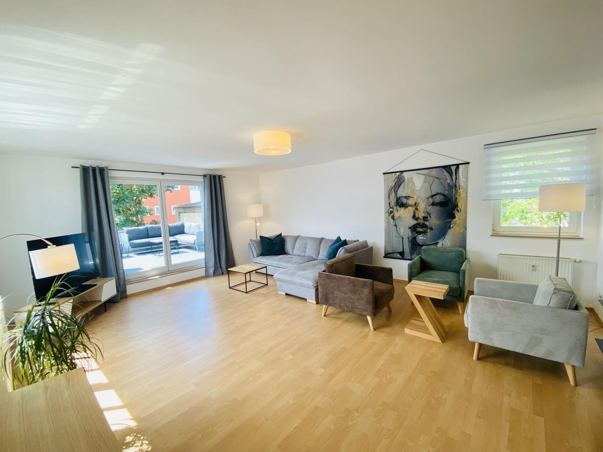 Wohnzimmer mit Sofa, Fernseher und Holzboden. Große Fenster und moderne Einrichtung.