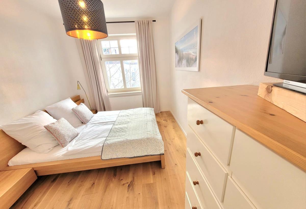 Schlafzimmer mit Bett, Fenster und Holzboden. An der Wand hängt ein Fernseher.