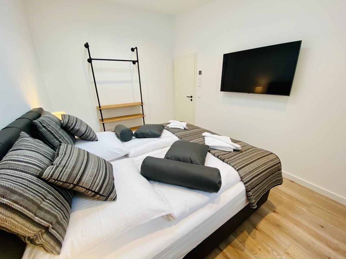 Schlafzimmer mit Bett, Nachttisch, Lampe und Wandfernseher. Holzboden und Fenster.