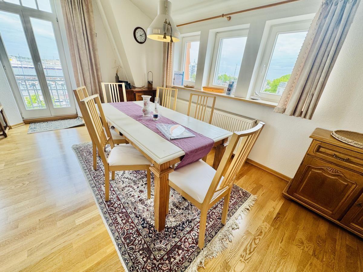 Esszimmer mit Tisch, Stühlen und Küche im Hintergrund. Holzboden und Fenster mit Vorhängen.