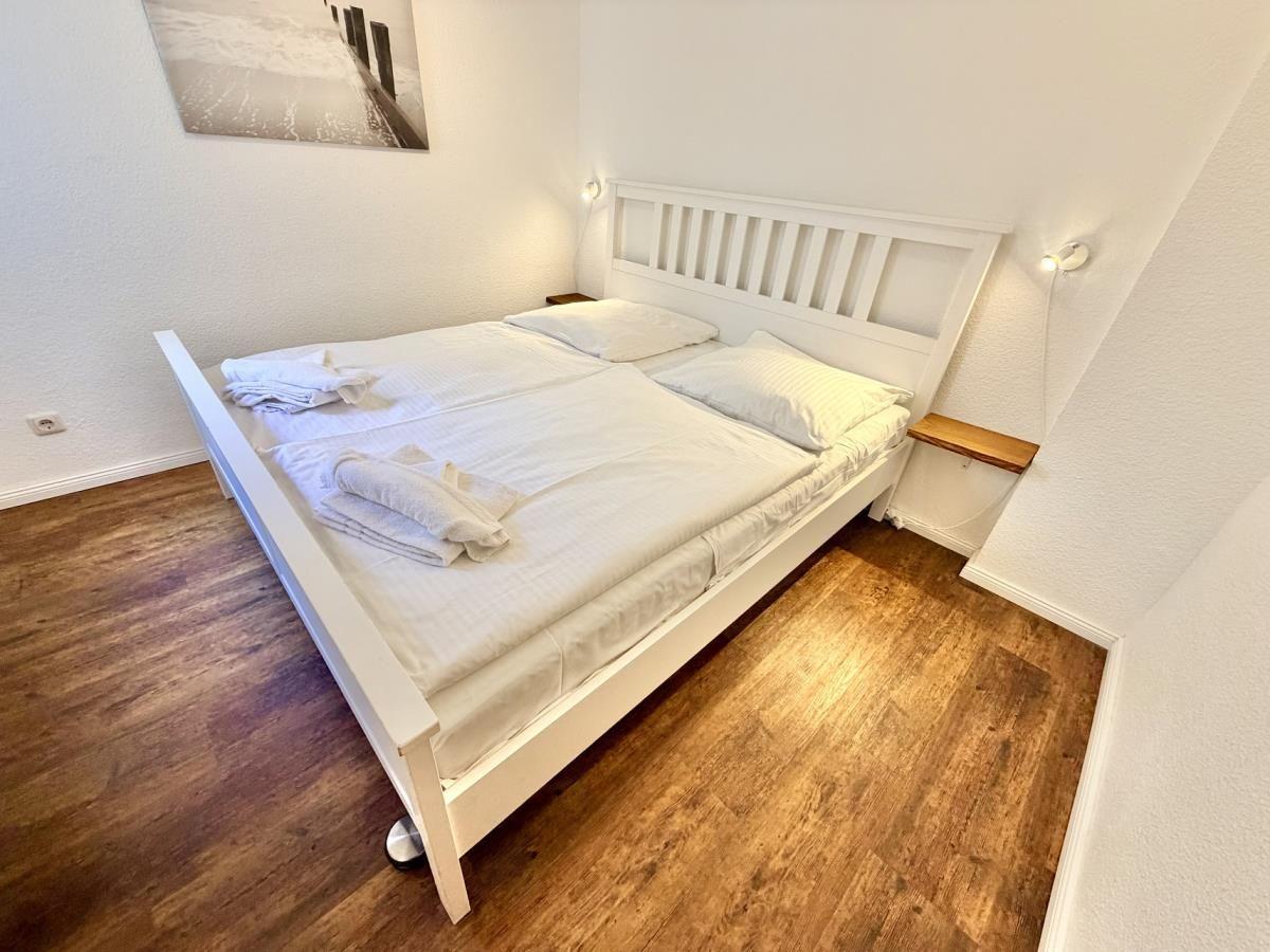 Schlafzimmer mit weißem Bett, Holzboden und Wandbild. Tür führt nach draußen.