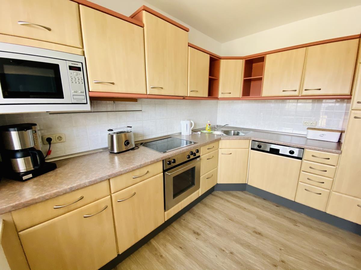 Küche mit Holzschränken, Esstisch und Fenster. Geräte wie Kühlschrank und Mikrowelle sind vorhanden.