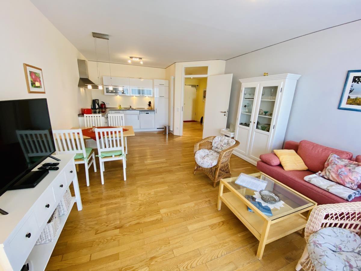 Wohnzimmer mit Sofa, Stühlen, Fernseher und Balkon. Holzboden und Fenster mit Vorhängen.
