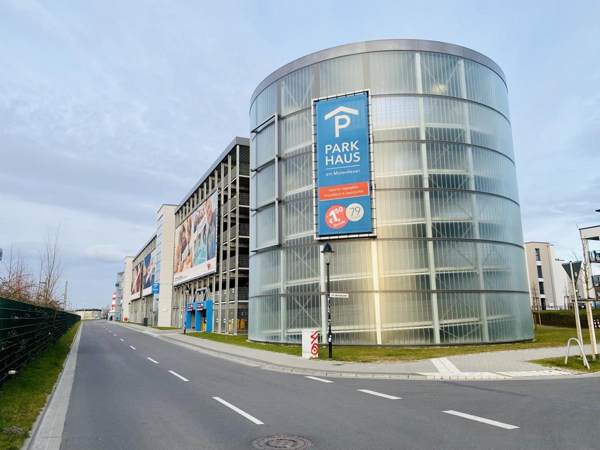 Multistock-Parkhaus mit Werbebanner und Eingang. Straße mit Grünflächen und Zaun.
