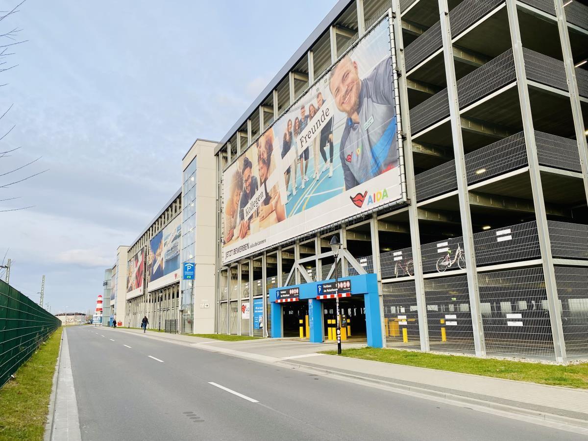 Parkhaus mit großen Werbeplakaten an modernem Gebäude. Straße mit Grünfläche und Zaun.