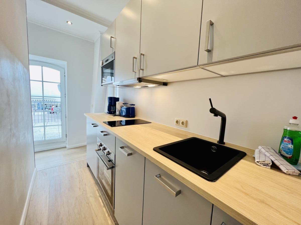 Küche mit Arbeitsplatte, Spüle und Dunstabzugshaube. Wohnzimmer mit Sofa und Fenster sichtbar.