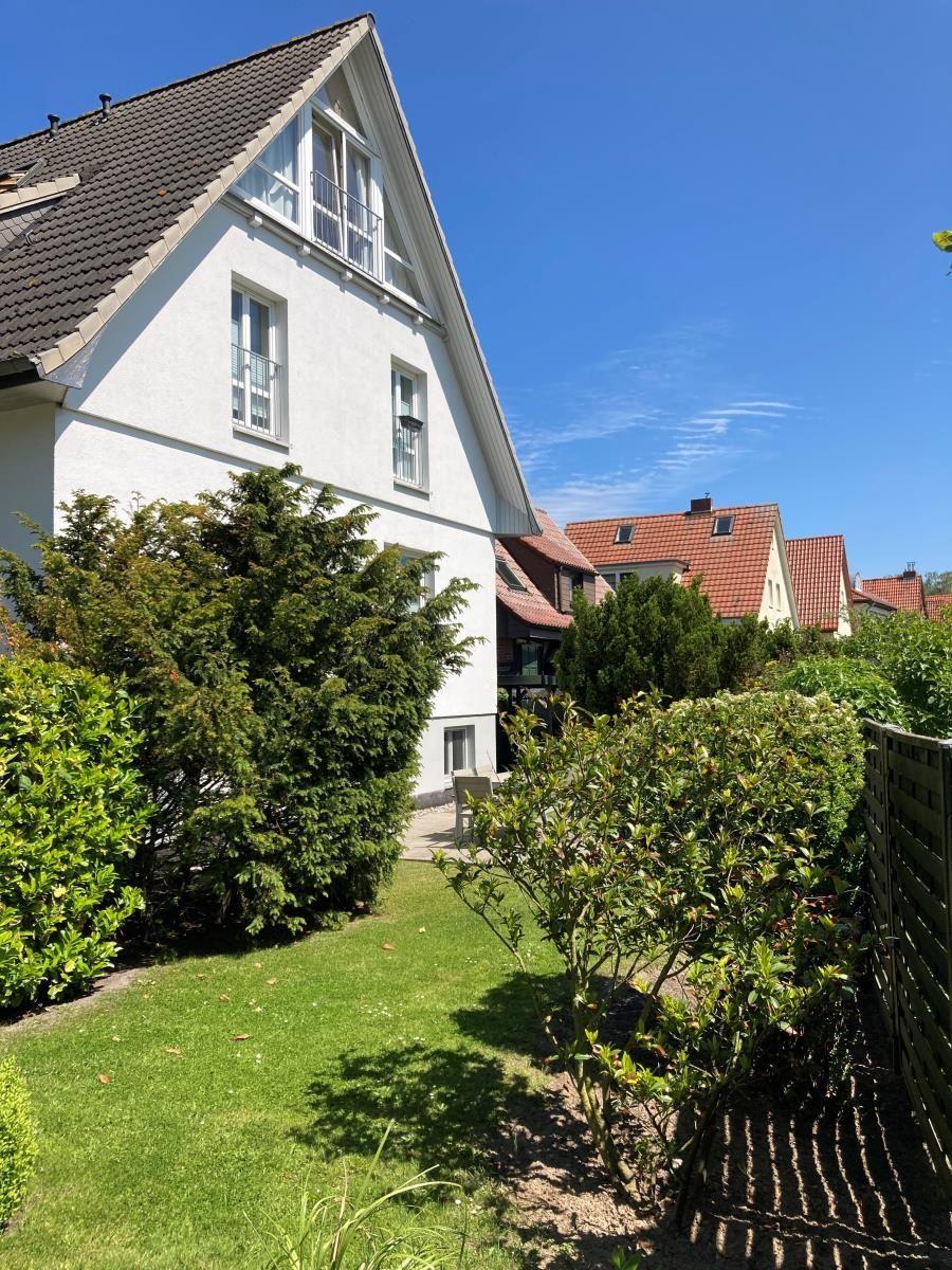 Ein zweistöckiges Haus mit dunklem Dach und weißer Fassade, umgeben von grüner Vegetation und einem Parkplatz.