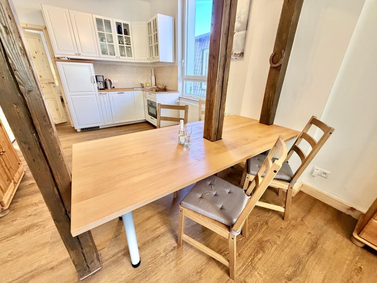 Essbereich mit Holztisch und Stühlen. Im Hintergrund Wohnzimmer mit Sofa und Fenster.