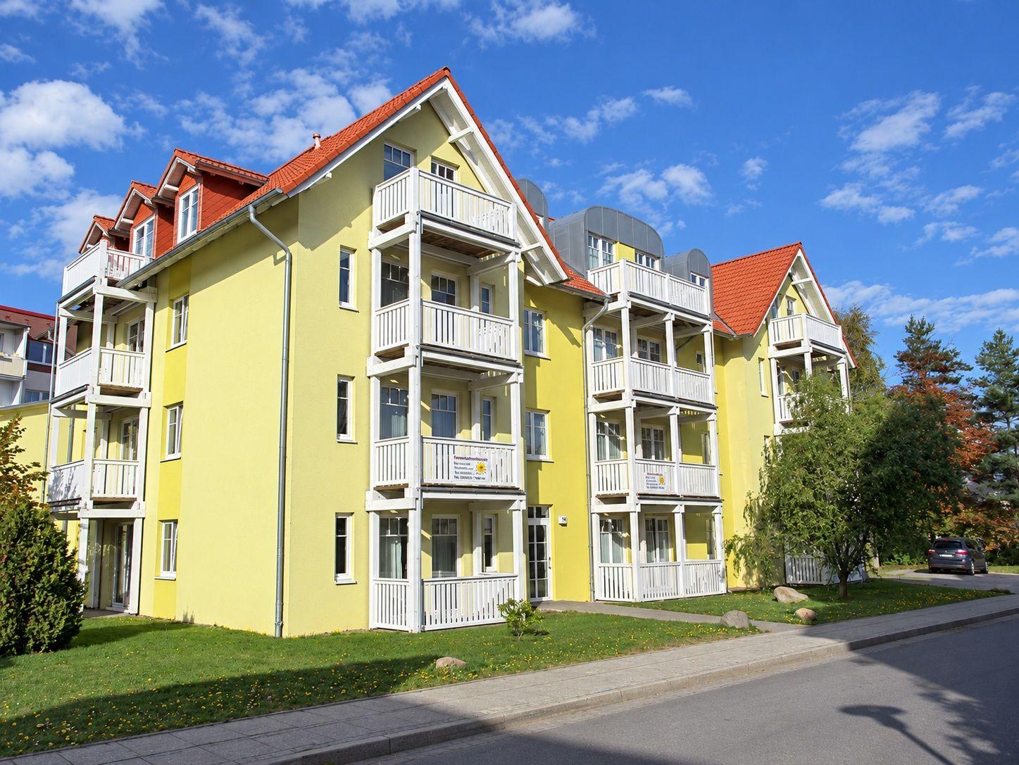 Gelbes Mehrfamilienhaus mit weißen Balkonen und rotem Dach unter blauem Himmel.